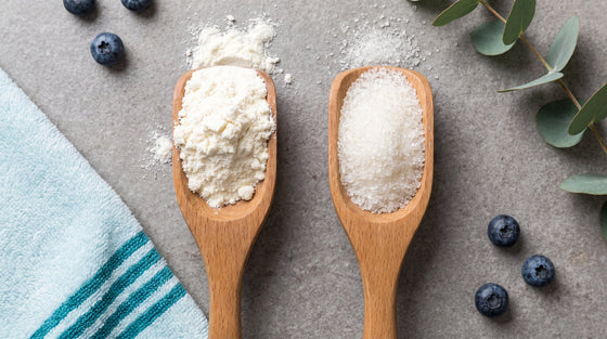 A whey protein powder scoop beside a collagen powder scoop on grey stone, comparing two popular fitness supplements.