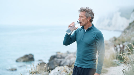 An active grey-haired man in his 50s drinking water outdoors on the coast after exercise, representing healthy aging.