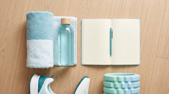 A post-workout recovery flat lay with a rolled towel, water bottle, notebook, sneakers, and foam roller on a light wood floor.