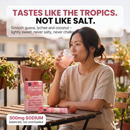 A woman sits at a wooden table on a balcony, drinking a pink beverage. A pink box labeled Sea Moss Hydration and a matching tube are placed on the table beside a small red fruit. morning glow