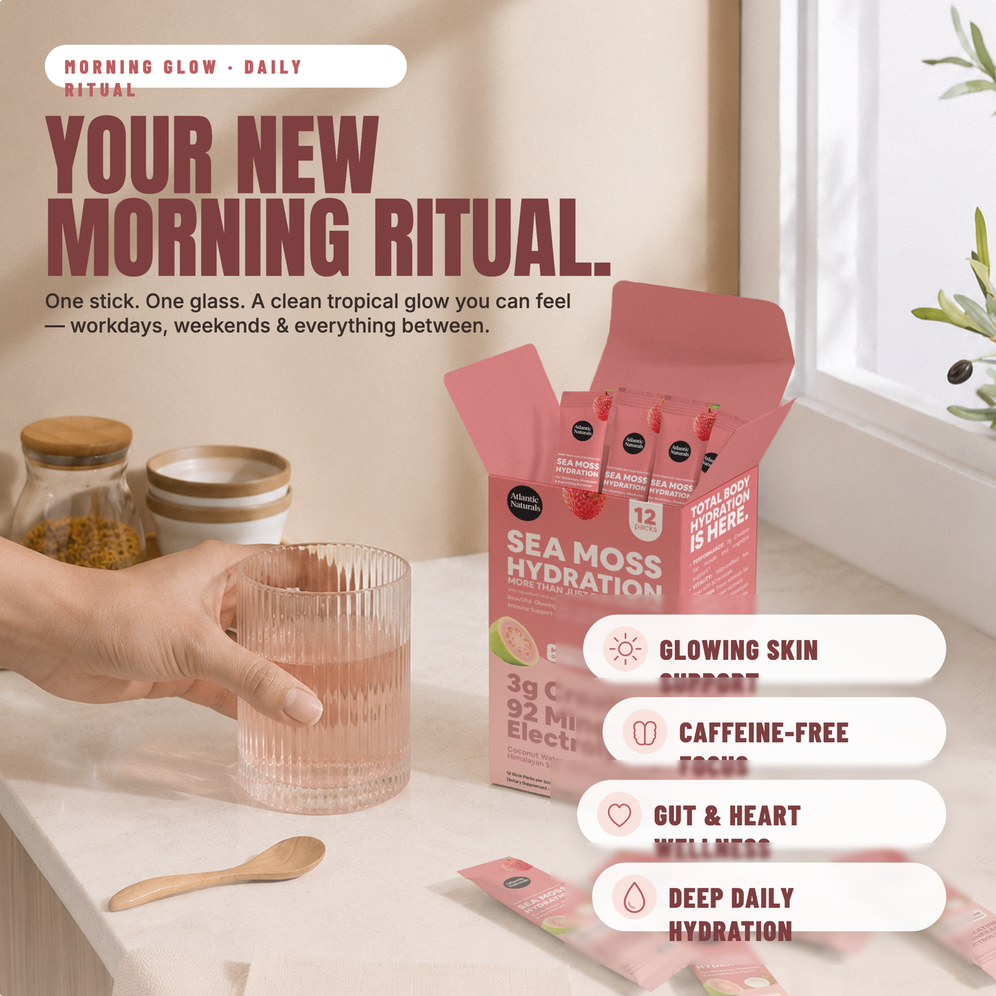 A hand holds a glass over a pink sea moss hydration powder box on a light countertop. The box is pink with black text and shows 12 individual stick packs inside. morning glow